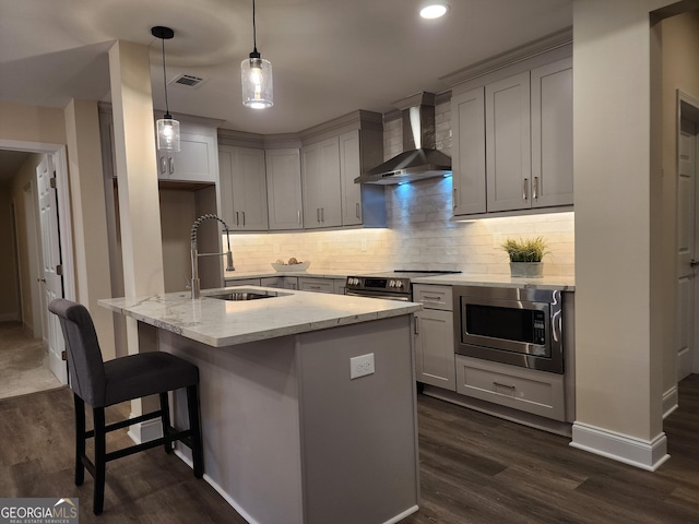 kitchen featuring light stone counters, wall chimney exhaust hood, stainless steel appliances, sink, and an island with sink