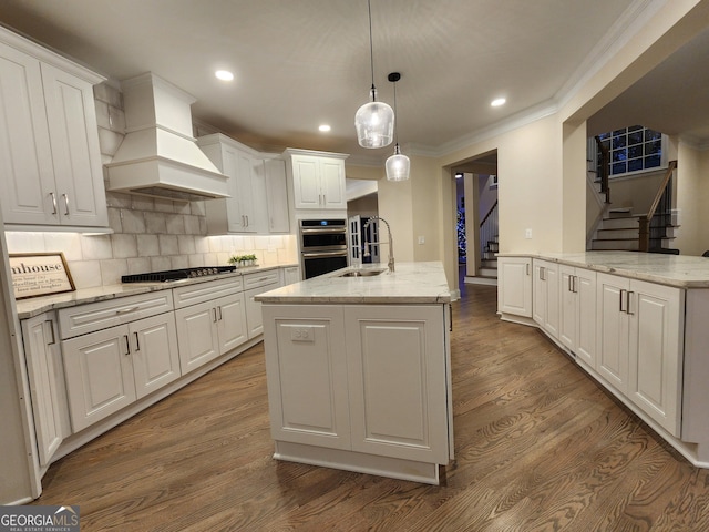 kitchen with appliances with stainless steel finishes, custom range hood, wood-type flooring, decorative light fixtures, and an island with sink