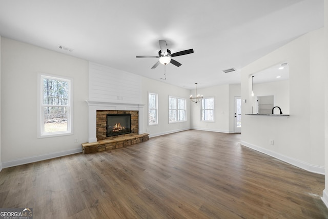 unfurnished living room featuring ceiling fan with notable chandelier, dark wood-type flooring, a stone fireplace, and a healthy amount of sunlight