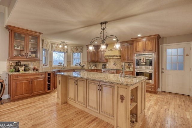 kitchen with an island with sink, light stone countertops, stainless steel double oven, sink, and decorative light fixtures