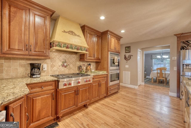 kitchen featuring stainless steel appliances, custom range hood, light wood-type flooring, tasteful backsplash, and a notable chandelier