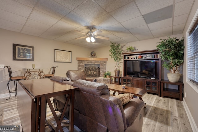 living room featuring a stone fireplace, light hardwood / wood-style floors, ceiling fan, a wealth of natural light, and a drop ceiling