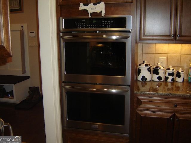 kitchen with dark brown cabinets, stainless steel double oven, tasteful backsplash, and dark stone countertops