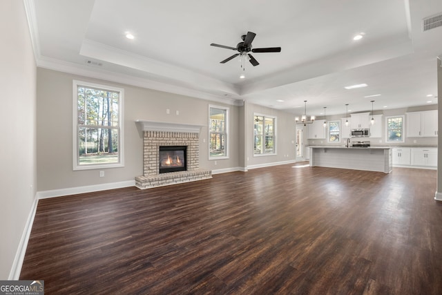 unfurnished living room with a tray ceiling, dark hardwood / wood-style flooring, ceiling fan with notable chandelier, and a brick fireplace