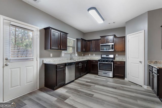 kitchen featuring stainless steel appliances, a sink, dark brown cabinetry, light stone countertops, and light wood-type flooring