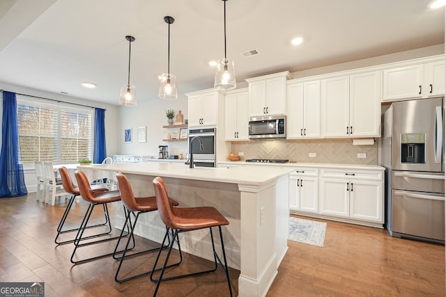 kitchen with white cabinets, stainless steel appliances, a center island with sink, and hanging light fixtures