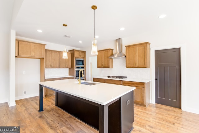 kitchen with hanging light fixtures, wall chimney range hood, an island with sink, appliances with stainless steel finishes, and light wood-type flooring