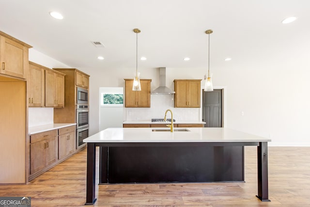 kitchen with an island with sink, pendant lighting, wall chimney range hood, and light wood-type flooring