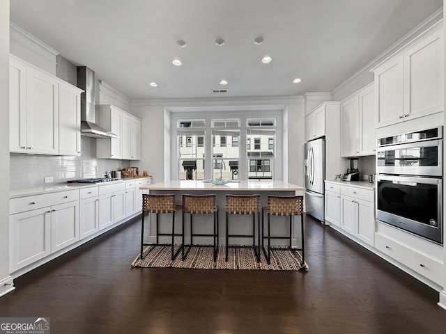 kitchen with white cabinetry, wall chimney range hood, an island with sink, and appliances with stainless steel finishes