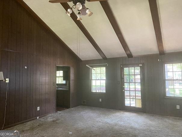 unfurnished living room with carpet flooring, vaulted ceiling with beams, ceiling fan, and wooden walls