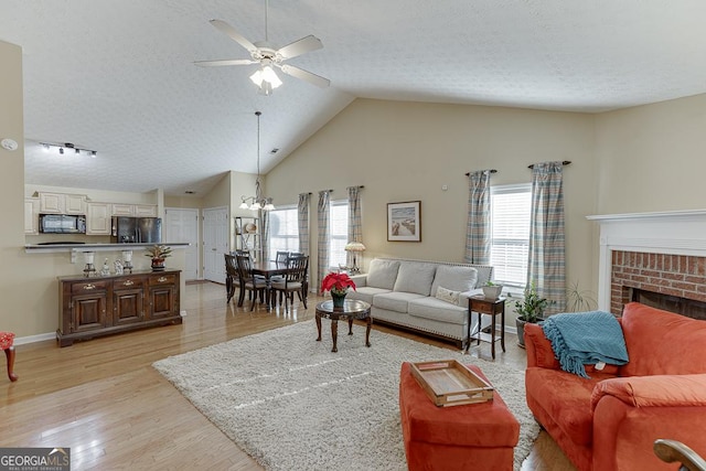 living room with a brick fireplace, high vaulted ceiling, ceiling fan with notable chandelier, and light wood-type flooring