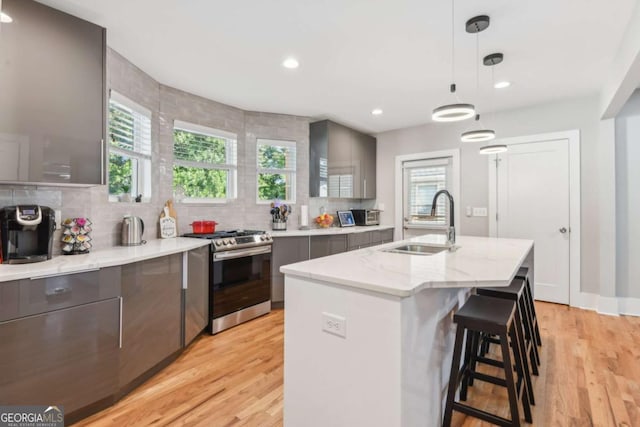 kitchen with sink, stainless steel range with gas stovetop, light hardwood / wood-style floors, hanging light fixtures, and an island with sink