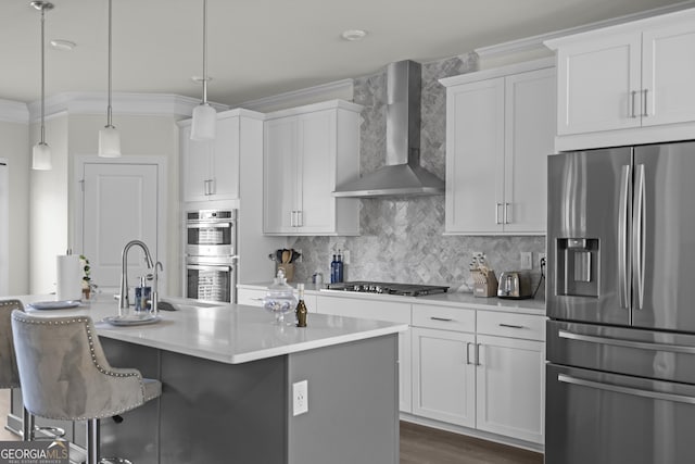 kitchen featuring appliances with stainless steel finishes, wall chimney exhaust hood, a center island with sink, white cabinetry, and hanging light fixtures