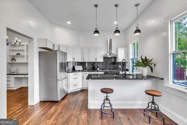 kitchen with white cabinets, wall chimney range hood, sink, kitchen peninsula, and stainless steel refrigerator