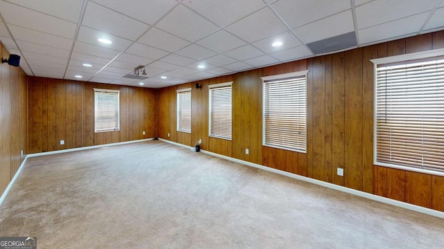 carpeted empty room featuring a drop ceiling, a healthy amount of sunlight, and wooden walls