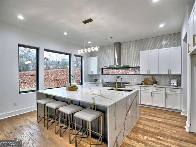 kitchen featuring white cabinetry, wall chimney exhaust hood, light stone counters, light hardwood / wood-style flooring, and a kitchen island with sink