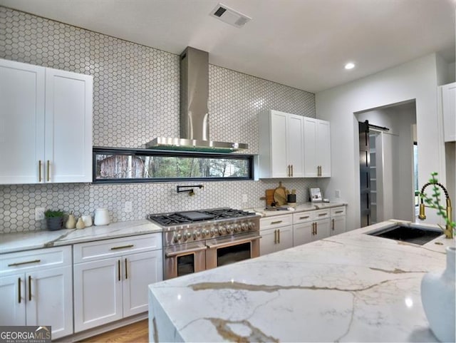 kitchen with light stone countertops, wall chimney exhaust hood, a barn door, high end stainless steel range, and white cabinets