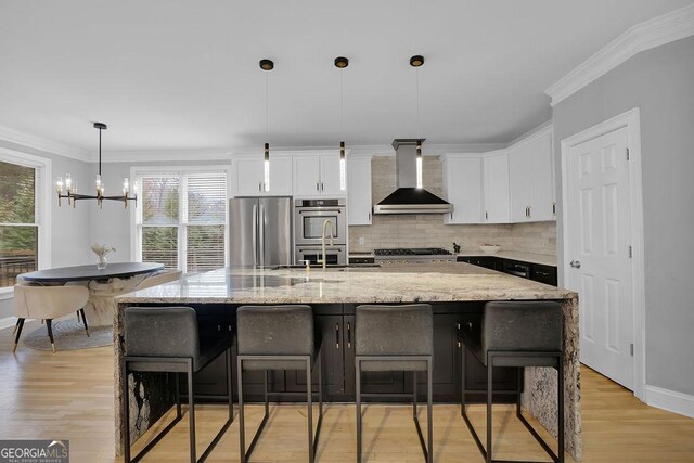 kitchen featuring light hardwood / wood-style flooring, wall chimney exhaust hood, appliances with stainless steel finishes, decorative light fixtures, and white cabinetry