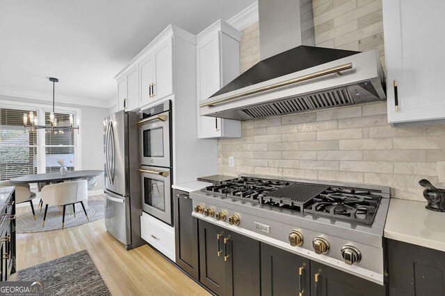 kitchen with appliances with stainless steel finishes, light wood-type flooring, wall chimney range hood, decorative light fixtures, and white cabinets