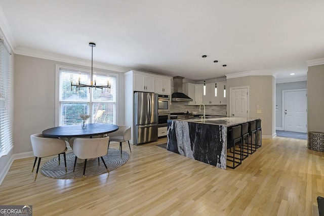 kitchen with appliances with stainless steel finishes, wall chimney exhaust hood, light hardwood / wood-style floors, white cabinetry, and hanging light fixtures