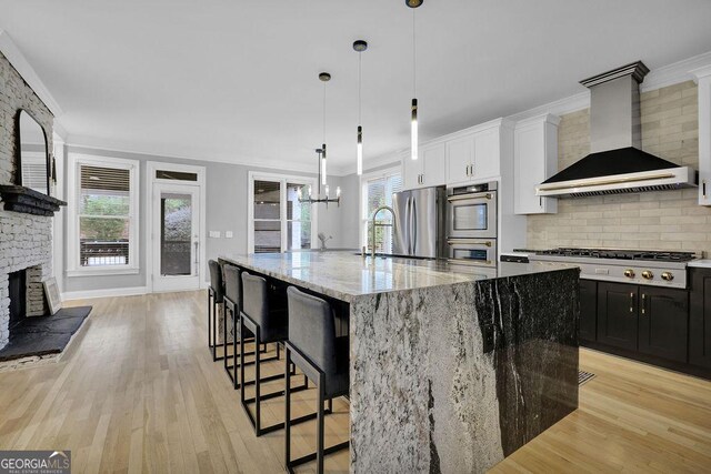 kitchen featuring a healthy amount of sunlight, a large island with sink, wall chimney exhaust hood, appliances with stainless steel finishes, and light hardwood / wood-style floors