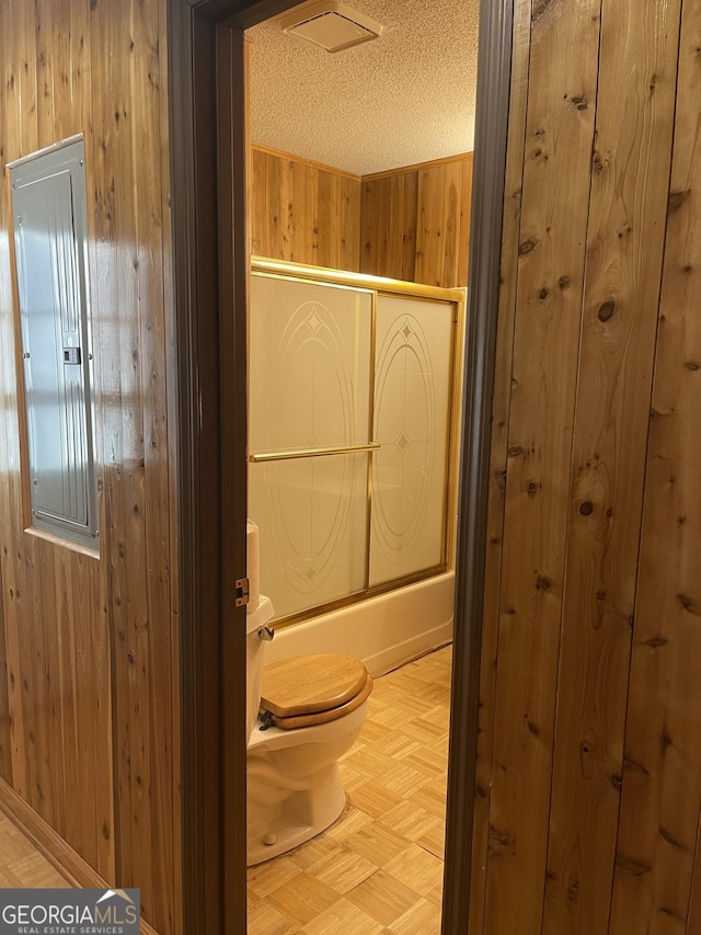 bathroom with parquet floors, electric panel, wood walls, a textured ceiling, and shower / bath combination with glass door