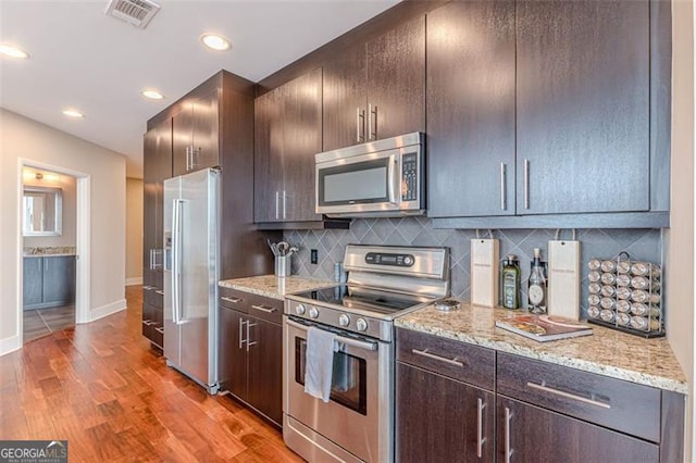 kitchen with backsplash, dark brown cabinets, light hardwood / wood-style floors, light stone counters, and stainless steel appliances