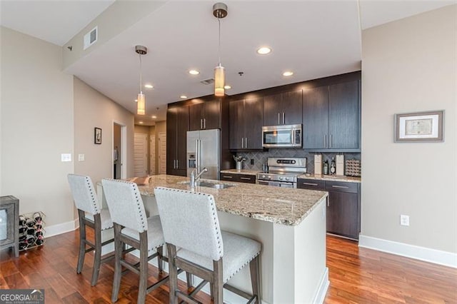 kitchen with stainless steel appliances, backsplash, pendant lighting, wood-type flooring, and a center island with sink