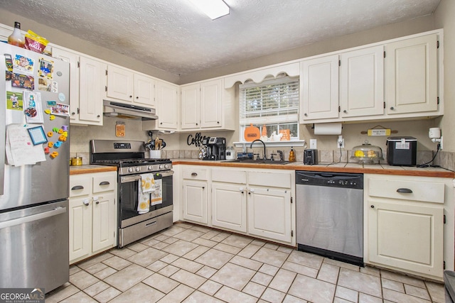 kitchen with under cabinet range hood, a sink, a textured ceiling, stainless steel appliances, and tile counters