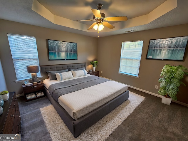 bedroom featuring a ceiling fan, baseboards, visible vents, a tray ceiling, and dark colored carpet