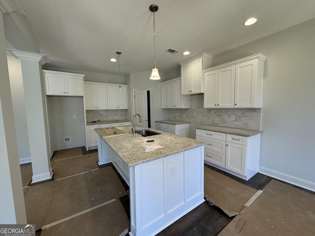 kitchen with light stone counters, a sink, white cabinets, hanging light fixtures, and an island with sink