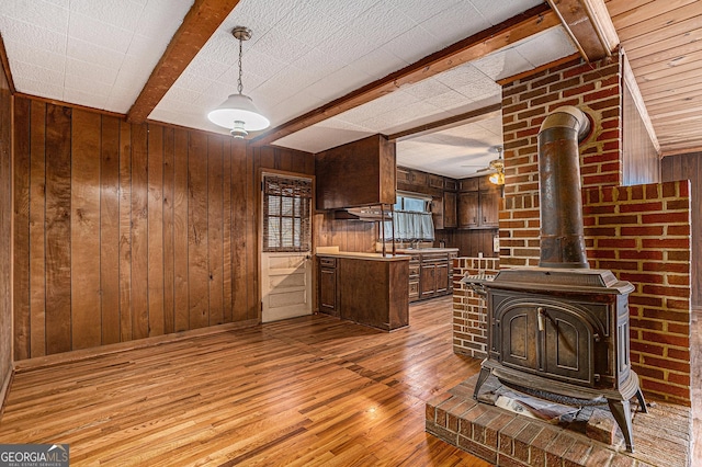 kitchen with beam ceiling, a wood stove, ceiling fan, light hardwood / wood-style floors, and wooden walls