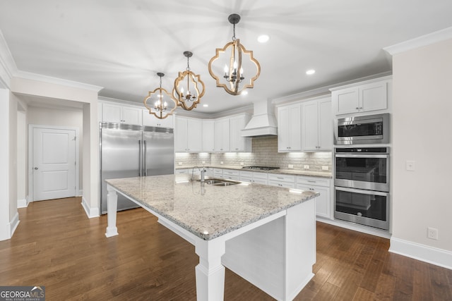 kitchen with dark hardwood / wood-style flooring, premium range hood, built in appliances, pendant lighting, and a kitchen island with sink