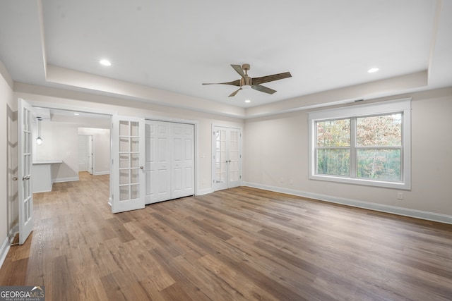 unfurnished bedroom featuring french doors, a raised ceiling, ceiling fan, wood-type flooring, and multiple closets