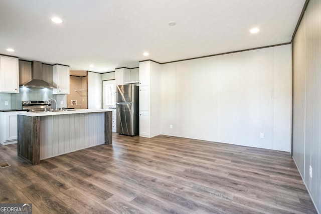 kitchen featuring white cabinets, an island with sink, stainless steel appliances, and wall chimney range hood