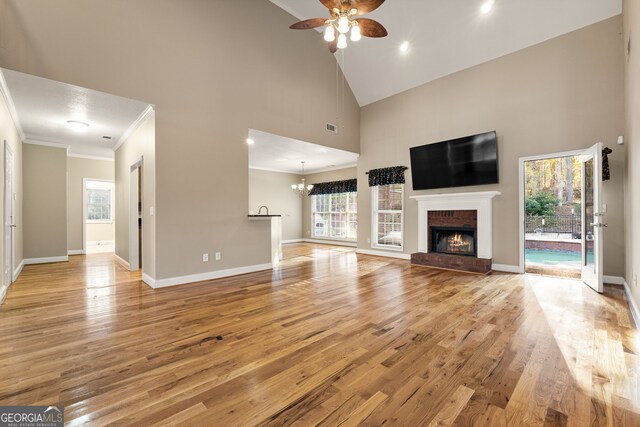 unfurnished living room featuring high vaulted ceiling, ceiling fan with notable chandelier, crown molding, a brick fireplace, and light hardwood / wood-style floors