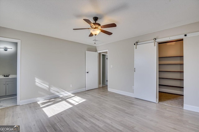 unfurnished bedroom with a walk in closet, ceiling fan, a barn door, connected bathroom, and light hardwood / wood-style floors