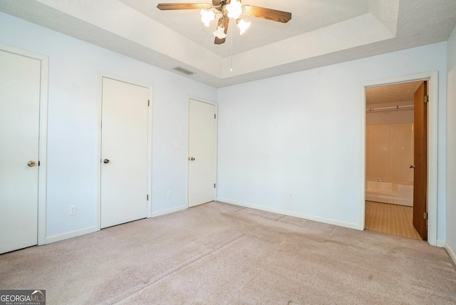 unfurnished bedroom featuring a raised ceiling, ensuite bath, ceiling fan, and light colored carpet