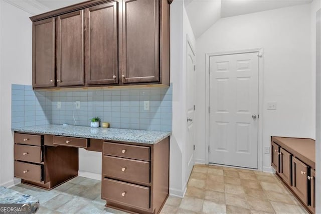 kitchen featuring backsplash, dark brown cabinetry, light stone counters, and built in desk