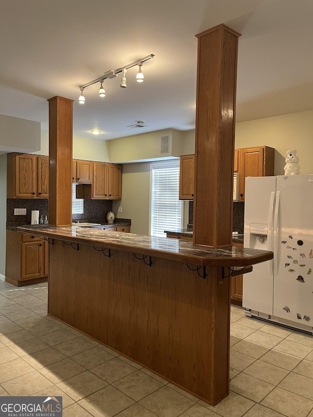 kitchen featuring light tile patterned flooring, backsplash, a kitchen breakfast bar, white fridge with ice dispenser, and kitchen peninsula