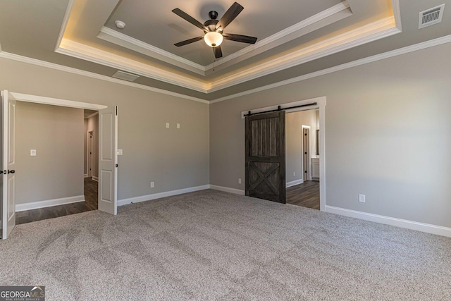 unfurnished bedroom with ceiling fan, a barn door, crown molding, a tray ceiling, and dark carpet