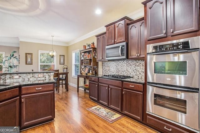 kitchen with appliances with stainless steel finishes, light wood-type flooring, dark stone counters, ornamental molding, and hanging light fixtures