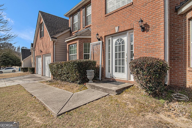 doorway to property featuring a garage, brick siding, driveway, and roof with shingles