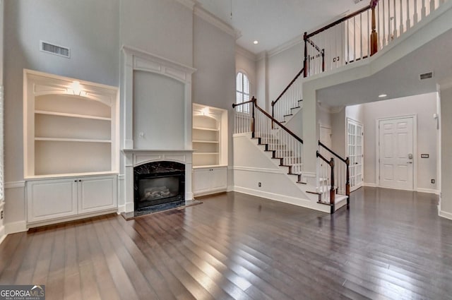 unfurnished living room featuring built in shelves, dark wood-type flooring, a premium fireplace, a towering ceiling, and ornamental molding