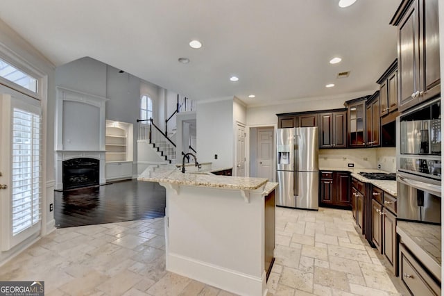 kitchen with a kitchen bar, a center island with sink, dark brown cabinets, light stone counters, and stainless steel appliances