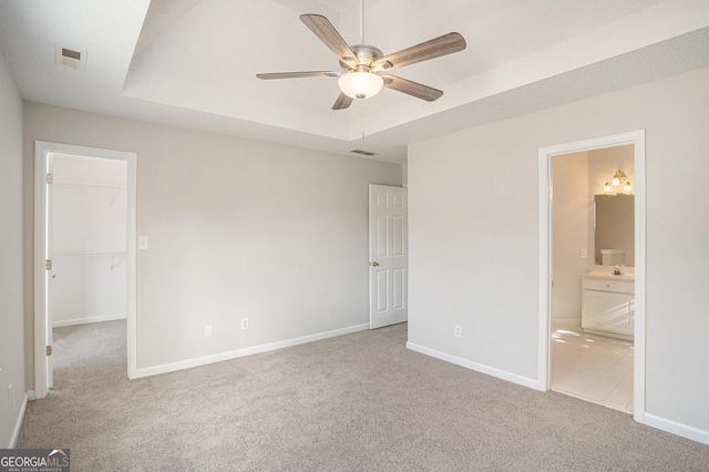 unfurnished bedroom with a tray ceiling, ensuite bath, ceiling fan, and light carpet