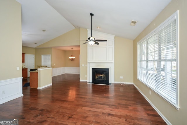 unfurnished living room with a large fireplace, a wainscoted wall, lofted ceiling, ceiling fan with notable chandelier, and dark wood-style flooring