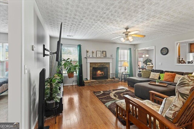 living room featuring hardwood / wood-style flooring, ceiling fan, a stone fireplace, and a textured ceiling