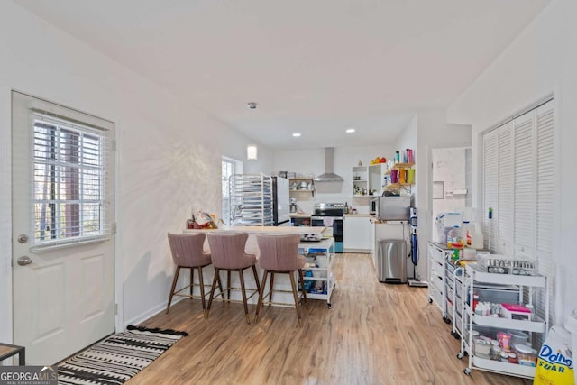 kitchen with stainless steel electric stove, wall chimney exhaust hood, light wood-type flooring, a kitchen bar, and white cabinetry