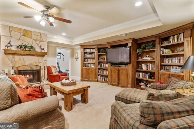 carpeted living room featuring a tray ceiling, a stone fireplace, ceiling fan, and ornamental molding